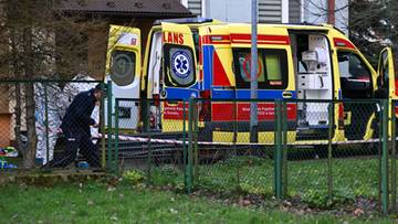 Karetka pogotowia ratunkowego z otwartymi drzwiami na tle budynku mieszkalnego, z widoczną osobą obok pojazdu i ogrodzeniem na pierwszym planie.