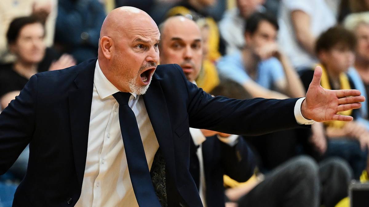 Mężczyzna w garniturze i krawacie instruuje z pasją na tle publiczności.