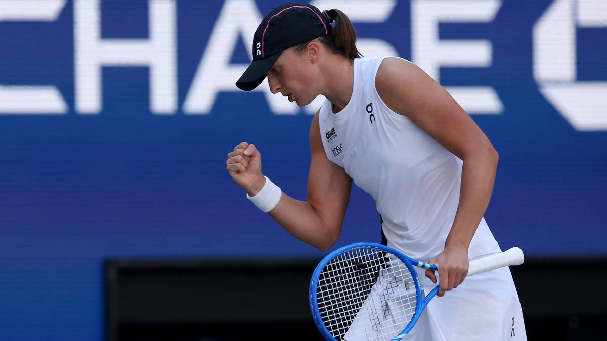 Iga Świątek, polska tenisistka, świętuje zdobycie punktu podczas meczu na US Open. Jest ubrana w białą koszulkę bez rękawów i czarną czapkę z daszkiem. W prawej ręce trzyma niebieską rakietę tenisową, a lewą pięść zaciska w geście radości.