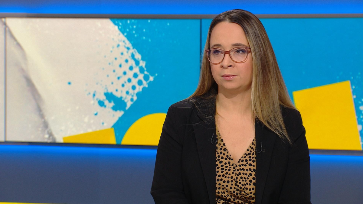 Kobieta w okularach i marynarce siedzi przed jasnym tłem z geometrycznymi wzorami.