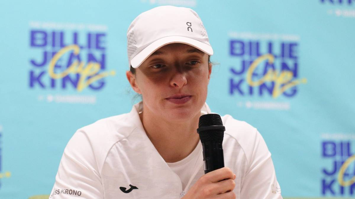 Iga Świątek w czapce i białej bluzie z mikrofonem w dłoni siedzi przy stole.