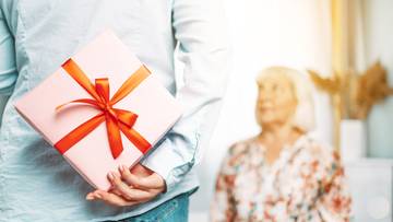 Osoba ukrywająca za plecami różowy prezent z czerwoną wstążką, w tle niewyraźna starsza kobieta.
