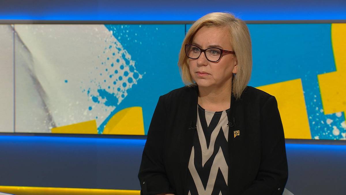 Kobieta w okularach i czarnej marynarce siedzi przed ekranem telewizyjnym z kolorowym abstrakcyjnym wzorem.