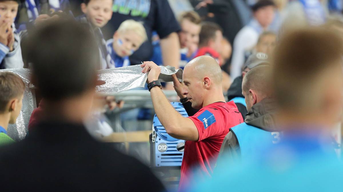 Sędzia piłkarski w czerwonej koszulce trzyma w górze materiał podobny do siatki do piłki siatkowej, podczas gdy wokół niego widać widzów na trybunach.