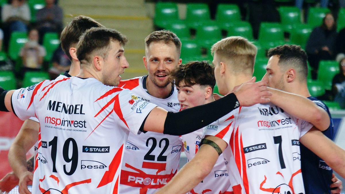 Grupa siatkarzy w białych strojach z czerwonymi elementami, przytulająca się w geście radości lub wsparcia. Na koszulkach widoczne są numery i logotypy sponsorów.