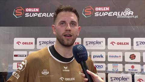 Kamil Adamski, siatkarz, udziela wywiadu z mikrofonem w ręku, na tle sponsor&oacute;w ligi siatk&oacute;wki.