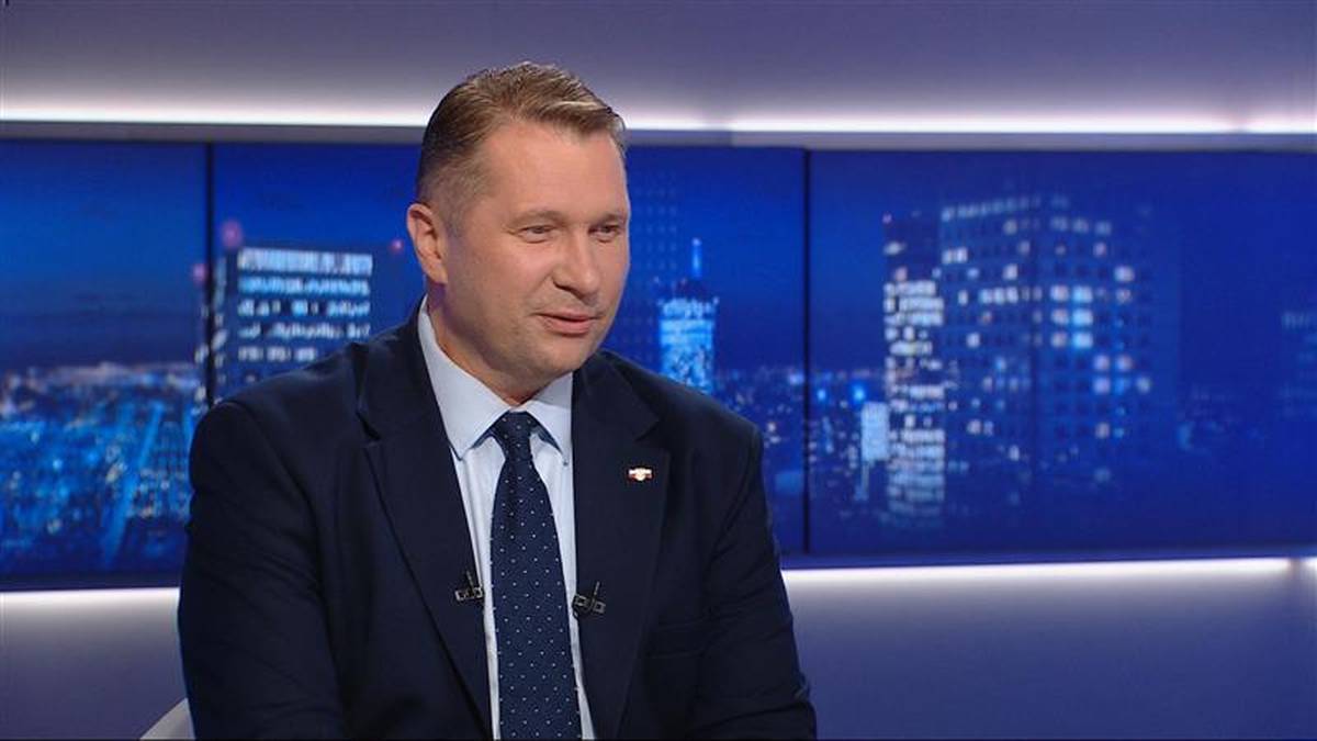 Mężczyzna w ciemnym garniturze i granatowym krawacie siedzi, patrząc w bok. W tle znajdują się trzy ekrany z nocnym widokiem miasta, oświetlonym licznymi budynkami.