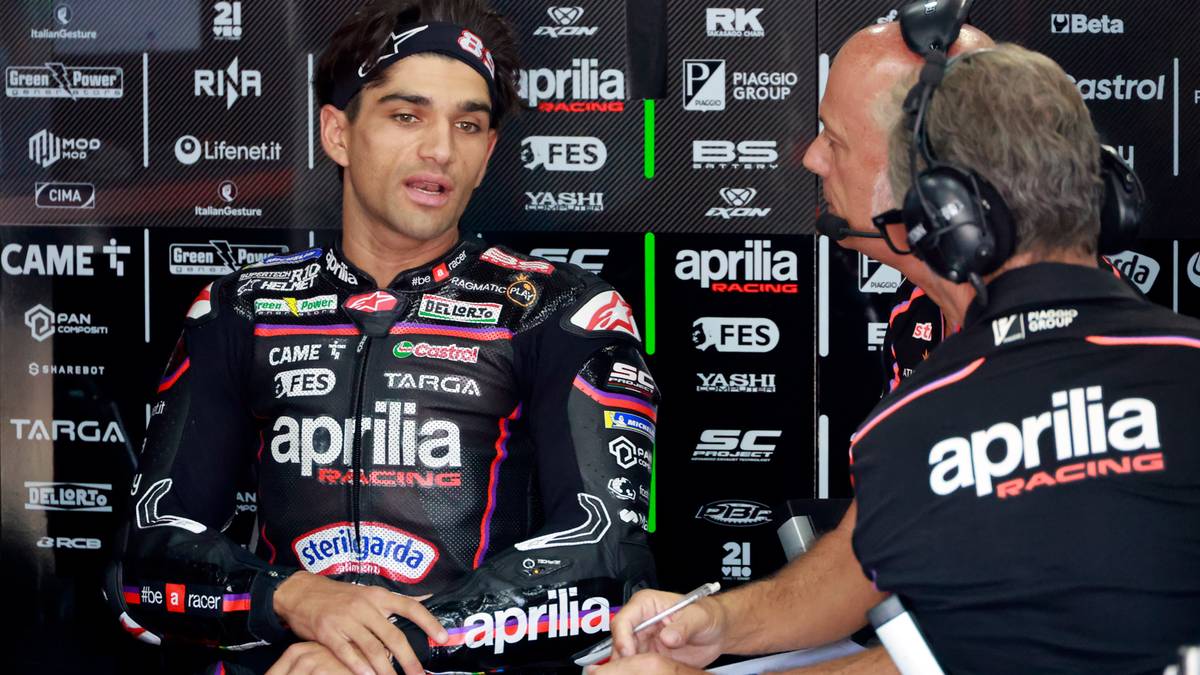 Dwóch mężczyzn w strojach wyścigowych. Jeden z nich, ubrany w czarną kurtkę wyścigową z logo Aprilia, ma opaskę na głowie i rozmawia z drugim mężczyzną siedzącym obok niego i trzymającym długopis.