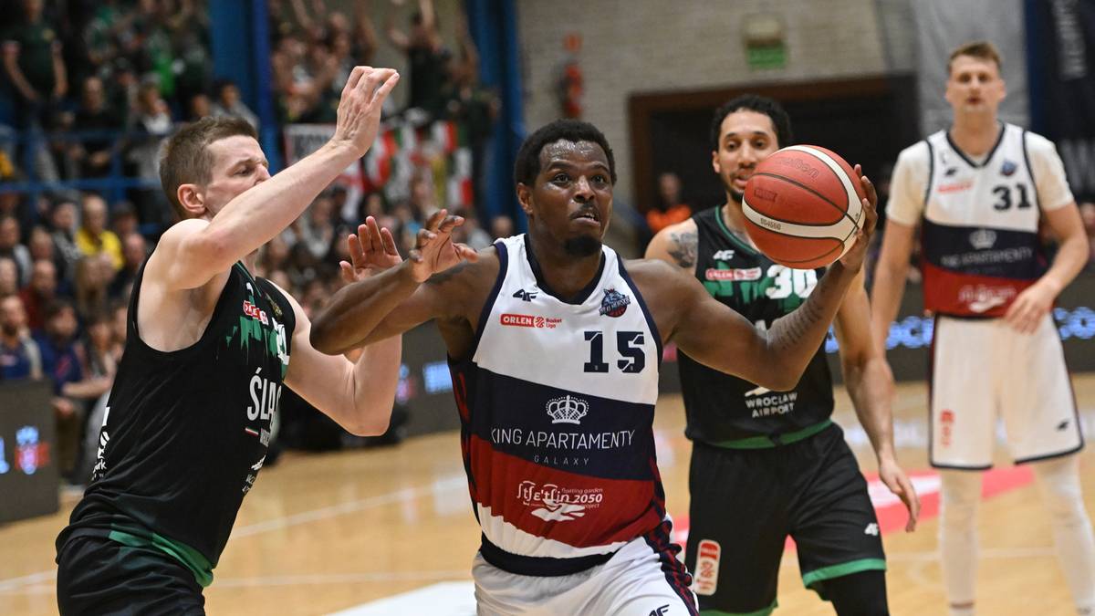 Koszykarz w czerwono-biało-granatowym stroju z numerem 15 na plecach, z piłką w rękach, podczas meczu. Dwóch obrońców z drużyny przeciwnej w czarnych strojach próbuje go zatrzymać.