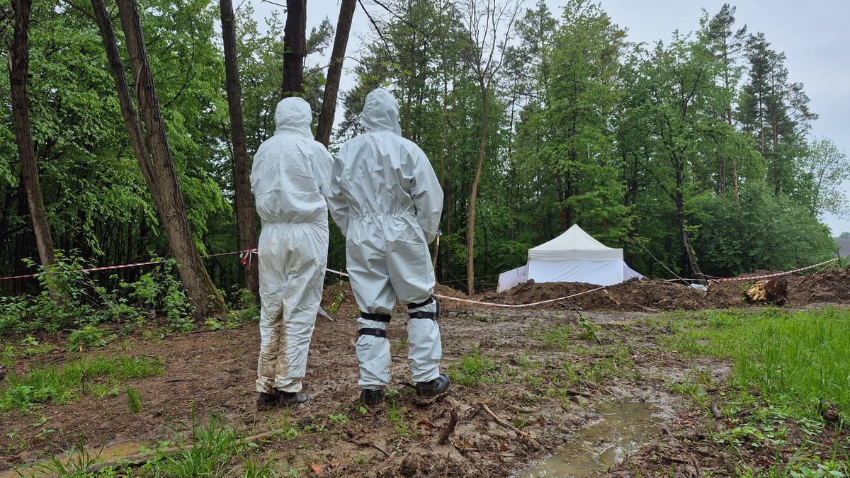 Dwie osoby w kombinezonach ochronnych stoją na błotnistym terenie z widocznym w tle namiotem.
