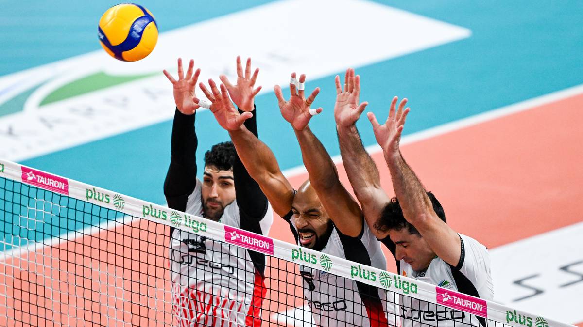 Czterech siatkarzy w białych i czerwonych strojach sportowych w skoku przy siatce, pr&oacute;bujących zablokować piłkę. Jeden z zawodnik&oacute;w na pierwszym planie podnosi ręce do g&oacute;ry, pozostałych trzech znajduje się za nim.