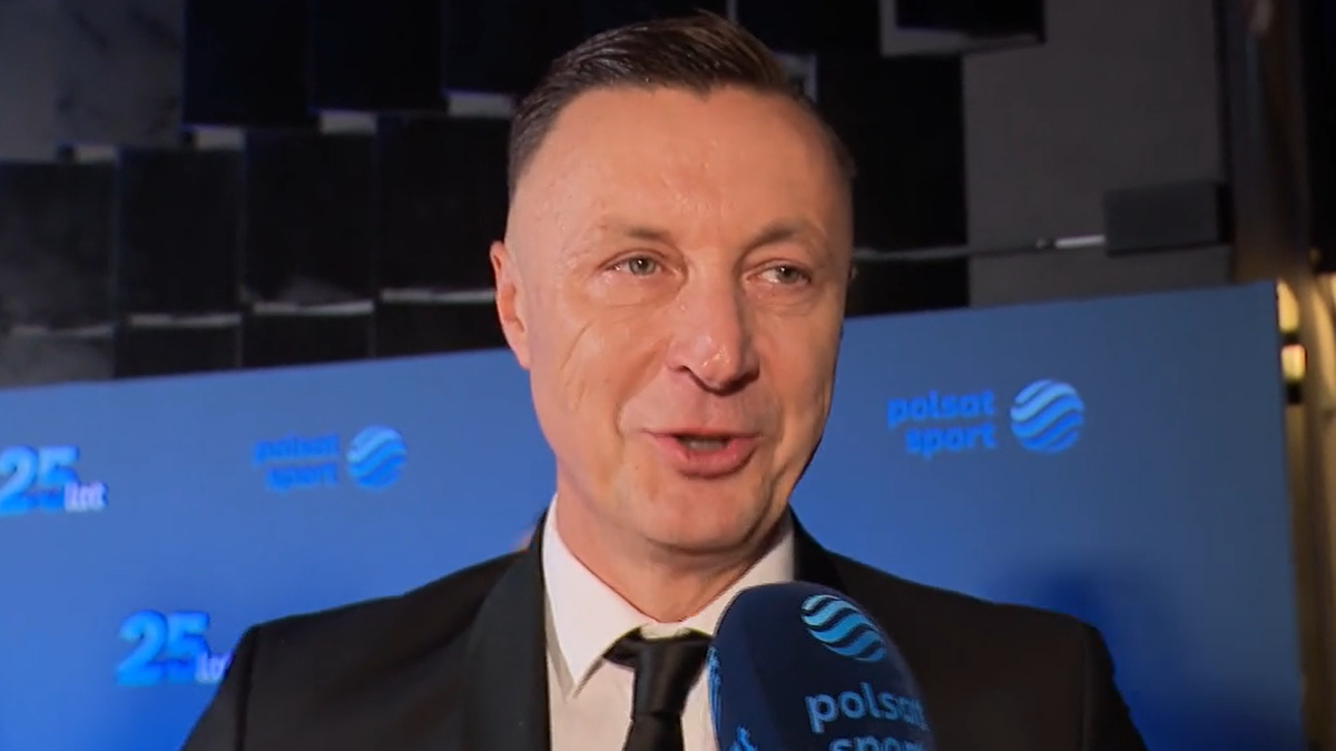 Tomasz Hajto w czarnym garniturze i krawacie, trzymający mikrofon Polsat Sport, podczas wywiadu na tle niebieskiego banera z logo stacji i napisem "25 lat".