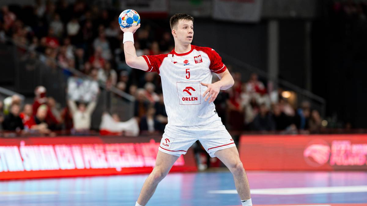 Szczypiornista w białej koszulce z czerwonymi elementami i numerem 5, przygotowuje się do rzutu piłką do piłki ręcznej na hali sportowej.