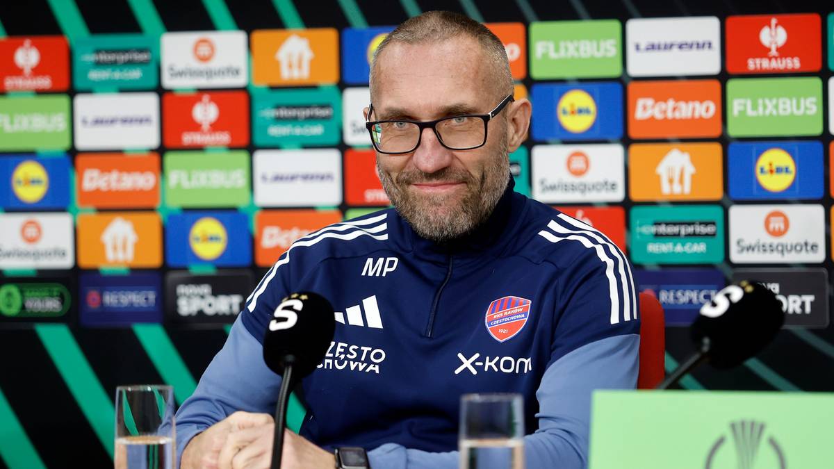 Trener siedzący przy stole podczas konferencji prasowej, ubrany w sportową kurtkę i okulary.