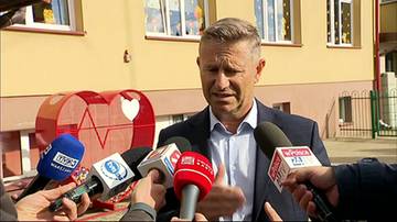 Mężczyzna w garniturze wypowiada się przed mikrofonami dziennikarzy na tle budynku szkoły.