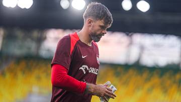 Mężczyzna w bordowej koszulce z czerwonymi rękawami, z kr&oacute;tkimi ciemnymi włosami, lekko pochylony, trzymający coś w rękach.