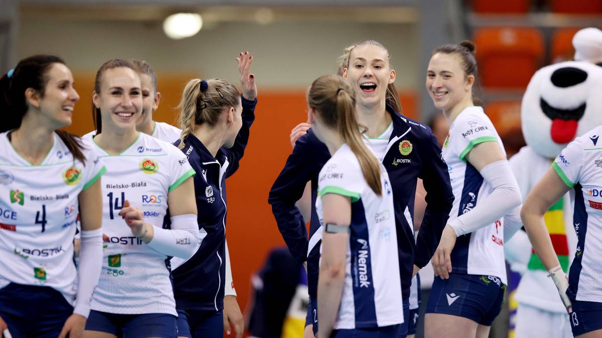 Grupa uśmiechniętych siatkarek w strojach sportowych na hali sportowej.