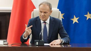 Donald Tusk siedzi przy biurku, wskazując palcem.