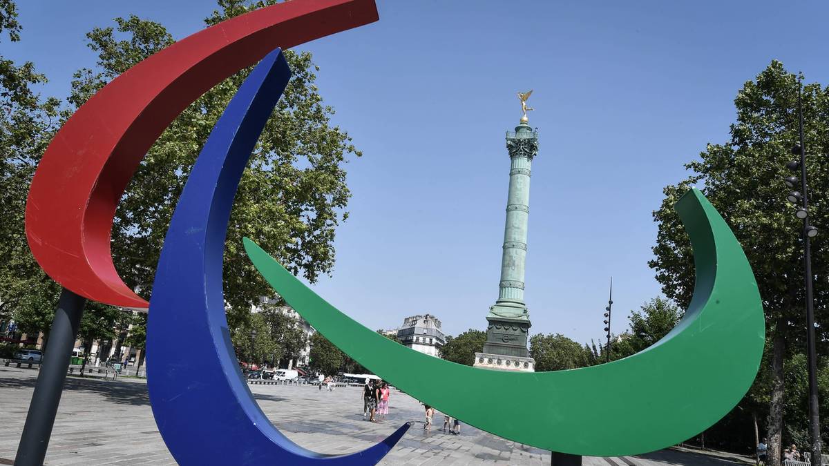 Trzy duże zakrzywione metalowe elementy w kolorach czerwonym, niebieskim i zielonym, tworzące abstrakcyjną rzeźbę, z kolumną na tle jasnego, błękitnego nieba.