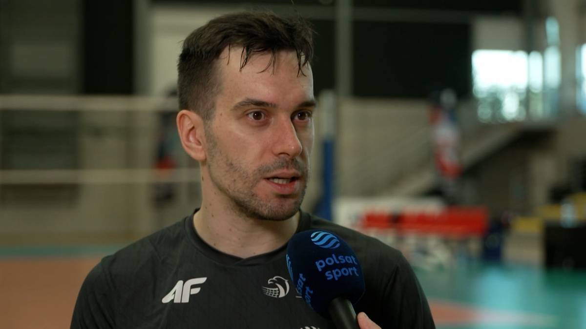 Mężczyzna w czarnej koszulce z logo "AF" i orłem, trzymający mikrofon z logo "Polsat Sport" podczas wywiadu w hali sportowej.