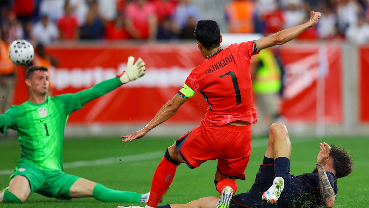 Bramkarz w zielonym stroju pr&oacute;buje obronić strzał piłkarza w czerwonym stroju, podczas gdy drugi zawodnik leży na murawie.