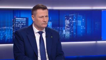 Krzysztof Paszyk siedzi na tle niebieskiego ekranu z grafiką przedstawiającą nocny widok miasta.