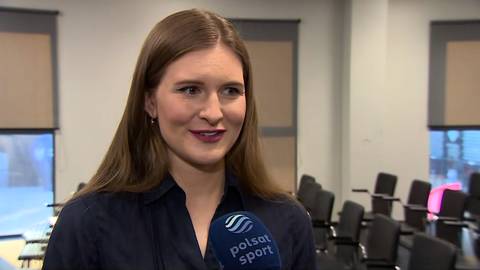 Portret kobiety z długimi brązowymi włosami, ubranej w ciemną koszulę, z mikrofonem Polsat Sport.