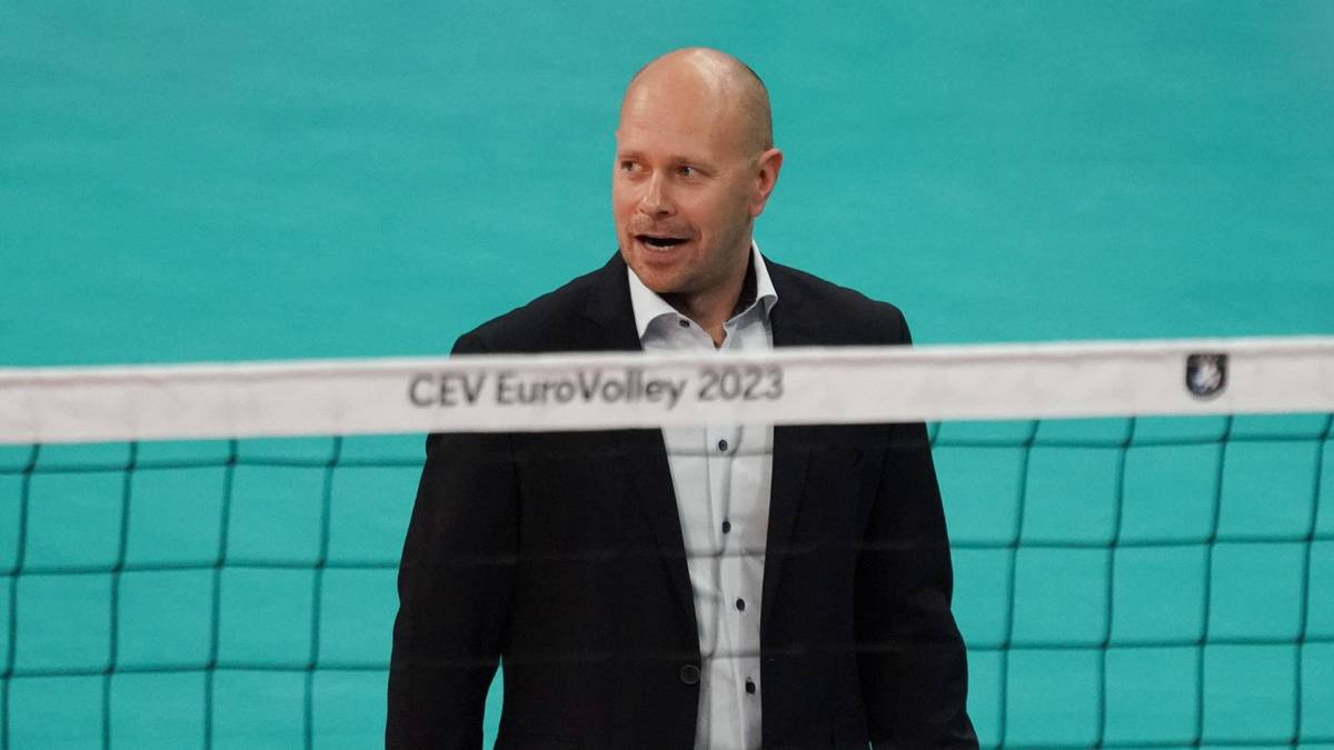 Łysy mężczyzna w garniturze z białą koszulą patrzy w bok z otwartymi ustami na tle siatki do siatkówki z napisem "CEV EuroVolley 2023".