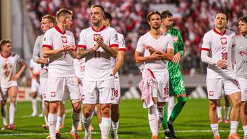 Piłkarze reprezentacji Polski w białych strojach stoją na boisku piłkarskim, w tle kibice.