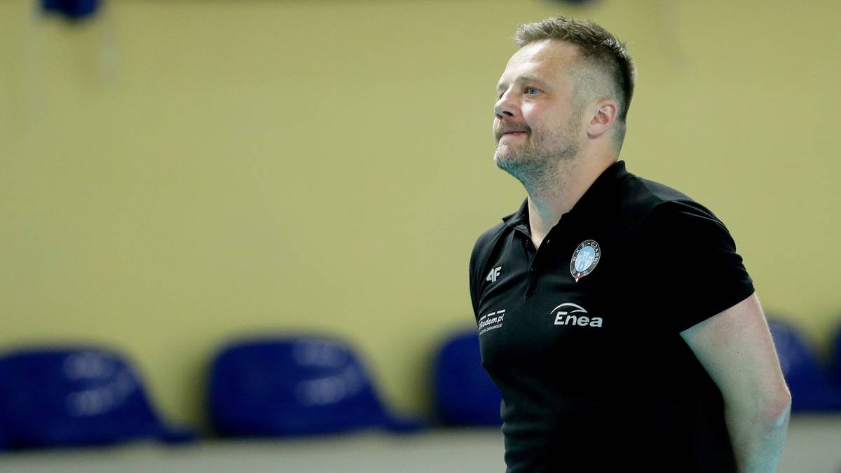 Mężczyzna w czarnej koszulce polo z logo Enea, lekko uśmiechnięty, patrzy w bok. Koszulka jest czarna, a w tle widoczne są żółta ściana i kilka niebieskich siedzeń.