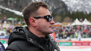 Trener skok&oacute;w narciarskich w okularach przeciwsłonecznych i czarnej kurtce zimowej, z widocznym fragmentem stadionu w tle.