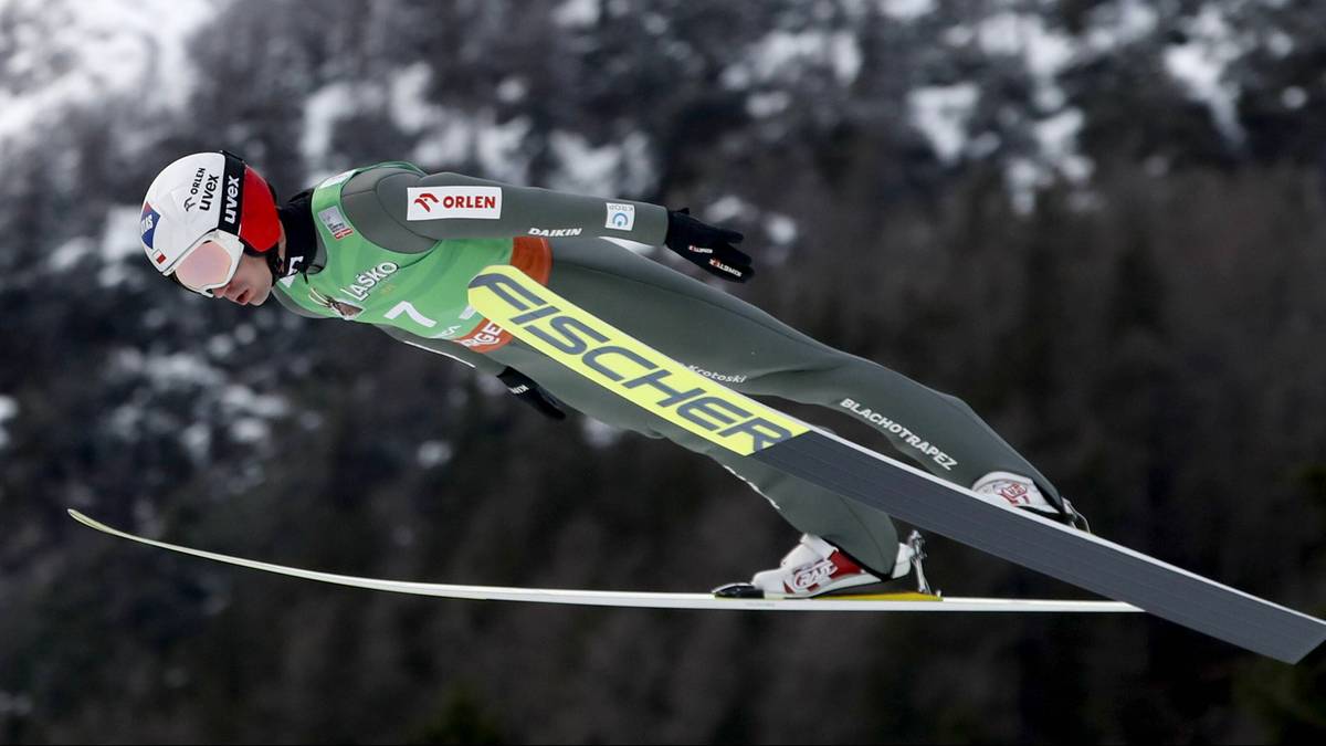 Skoczek narciarski w zielonym kombinezonie wykonuje skok, w tle widoczne są ośnieżone góry.