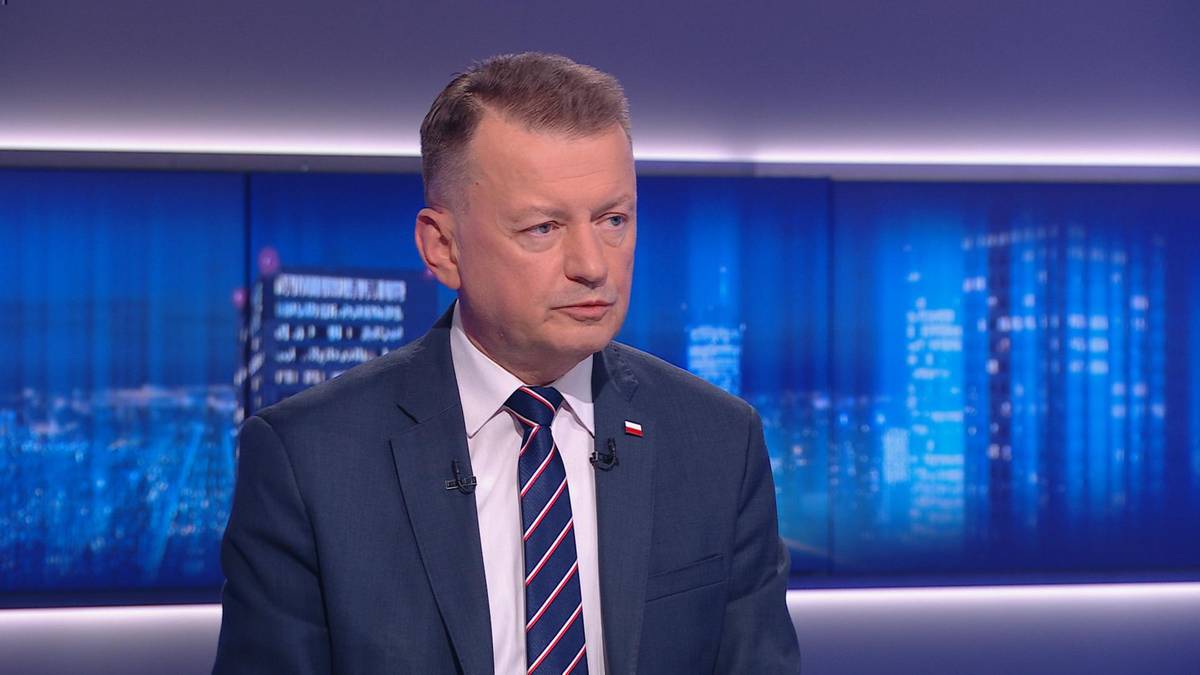 Mężczyzna w garniturze i krawacie siedzi na tle niebiesko-białego ekranu.