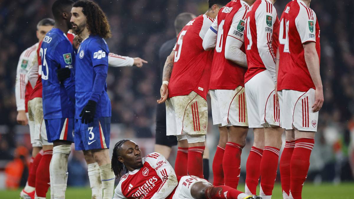 Piłkarz w czerwono-białym stroju Arsenalu leży na murawie, podczas gdy inni zawodnicy w czerwonych strojach stoją w pobliżu.