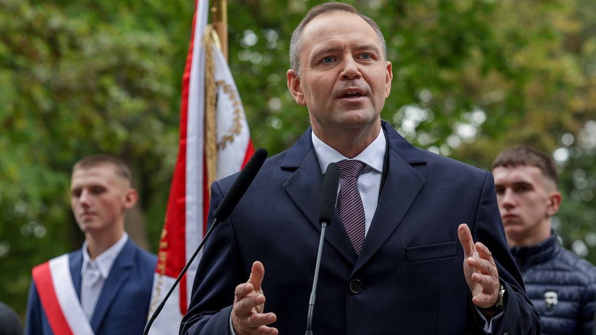 Mężczyzna w garniturze przemawia za mównicą. Za nim widoczne są dwie flagi, w tym polska, oraz rama z portretem.