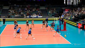 Siatkarki w niebieskich strojach rywalizują na boisku z pomarańczową nawierzchnią. Widownia jest wypełniona widzami.