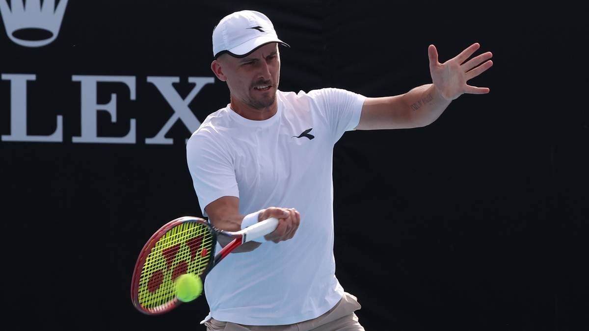 Tenisista w białej koszulce i czapce z daszkiem uderza piłkę rakietą podczas meczu. W tle logo Rolex.