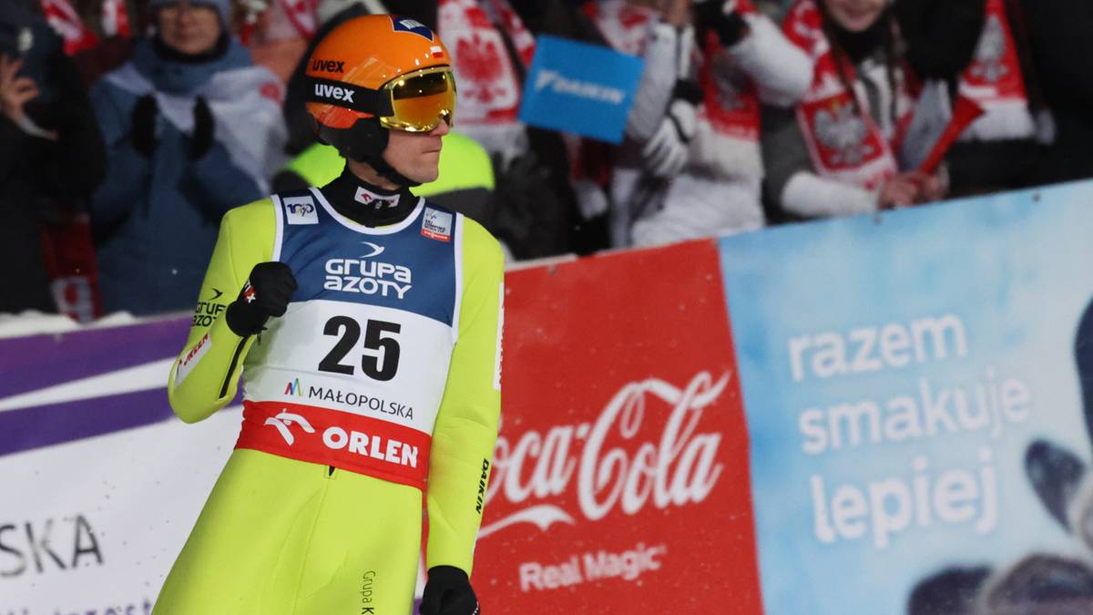 Skoczek narciarski w ż&oacute;łto-niebieskim kombinezonie z numerem 25 na piersi, przygotowujący się do skoku. Na tle widoczni są kibice i sponsorzy zawod&oacute;w.