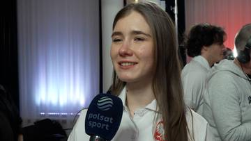 Młoda kobieta z długimi, rozpuszczonymi włosami, ubrana w białą koszulę, trzyma mikrofon z logo Polsat Sport i odpowiada na pytania. W tle widać inne osoby i oświetlenie.