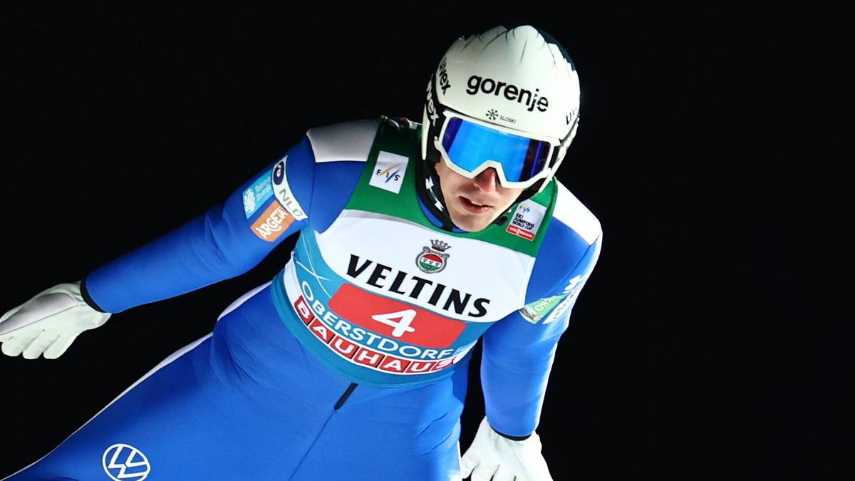 Skoczek narciarski w locie, ubrany w niebieski kombinezon i biały kask.