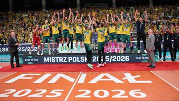 Drużyna siatkarzy w strojach sportowych wznosi ręce w geście radości na podium, na tle tłumu kibic&oacute;w. Na podium znajduje się napis "MISTRZ POLSKI 2025/2026".