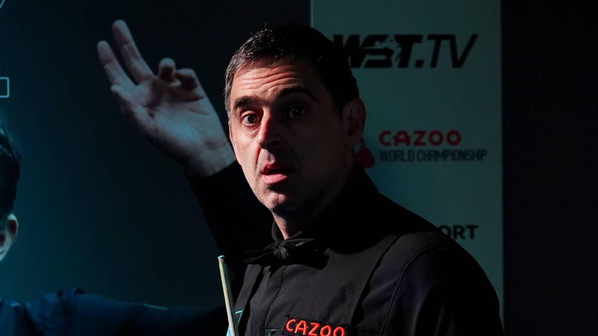 Ronnie O'Sullivan, znany snookerzysta, wskazuje palcem na bok, ubrany w czarną koszulę z muszką.
