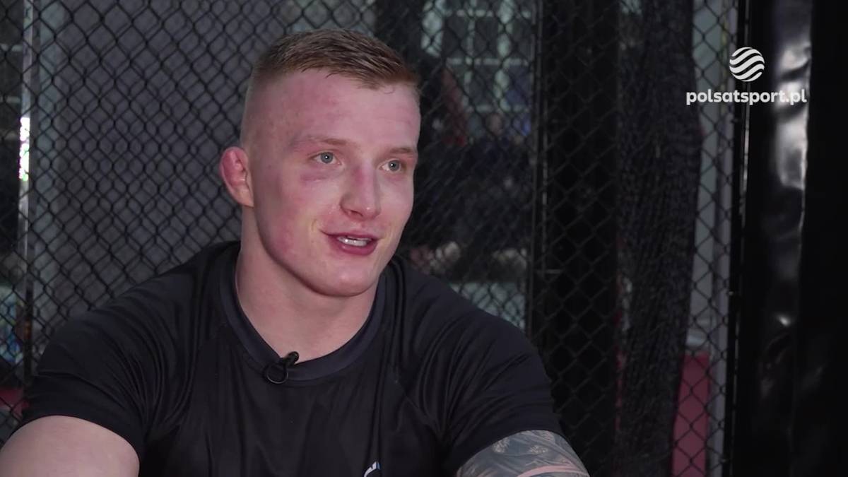 Młody mężczyzna o jasnej skórze i krótkich, jasnych włosach, ubrany w czarną koszulkę, siedzi przed siatką oktagonu.