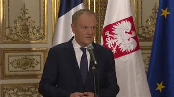 Donald Tusk przemawia przed mikrofonem, za nim widoczne są flagi Polski, Francji i Unii Europejskiej.