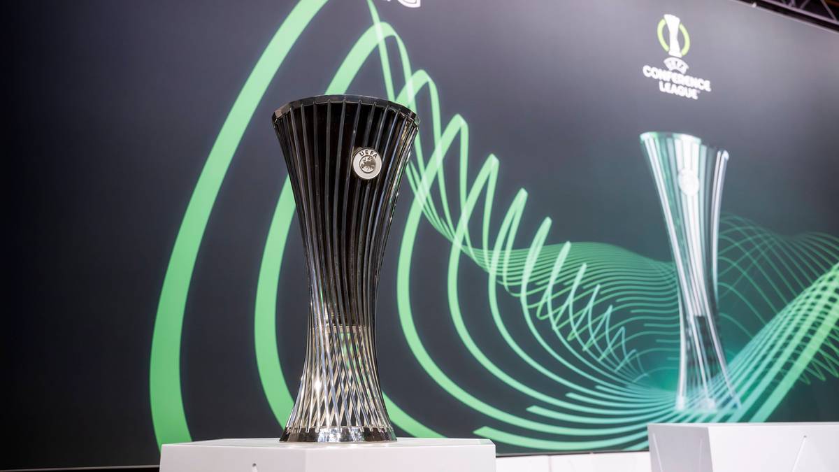 Dwa puchary piłkarskie, jeden duży i ciemny, drugi mniejszy i srebrny, na tle ściany z napisem "ROAD TO LEIPZIG".