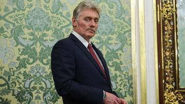 Dmitrij Pieskow w garniturze stoi na tle ściany z ozdobną tapetą.