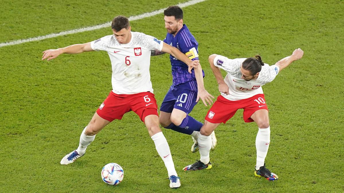 Trzech piłkarzy na zielonej murawie boiska piłkarskiego. Dwóch z nich w białych koszulkach z czerwonymi spodenkami i dwoma białymi numerami na koszulkach, trzeci w niebiesko-fioletowej koszulce z numerem 10. Dwóch białych graczy próbuje odebrać piłkę trzeciemu.