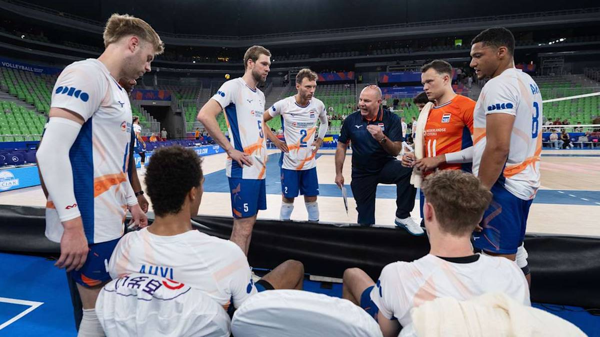 Drużyna siatkarzy w białych koszulkach z niebieskimi elementami, słucha trenera w niebieskiej koszulce podczas przerwy w meczu na hali sportowej z zielonymi trybunami w tle.