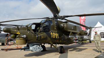 Zdjecie przedstawia śmigłowiec szturmowy Mi-28NE w ciemnozielonych barwach wojskowych, stojący na płycie lotniska. Na kadłubie widoczny jest numer "1811". W tle widać fragmenty hangar&oacute;w i inne obiekty lotnicze.