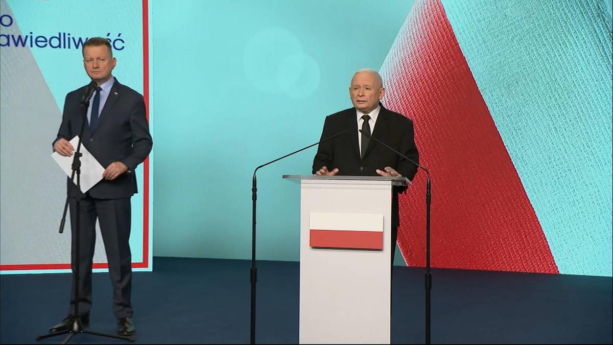 Dw&oacute;ch mężczyzn w garniturach stoi na scenie, jeden przemawia przy m&oacute;wnicy, drugi trzyma dokumenty.